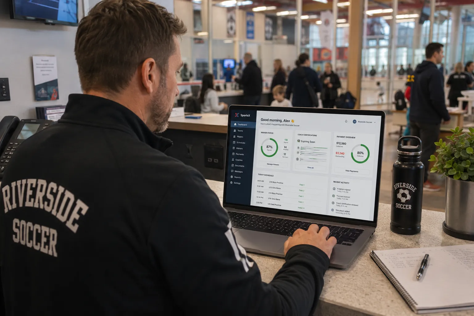 Club manager reviewing the SportsX morning dashboard on a laptop at the front desk of a busy community sports facility